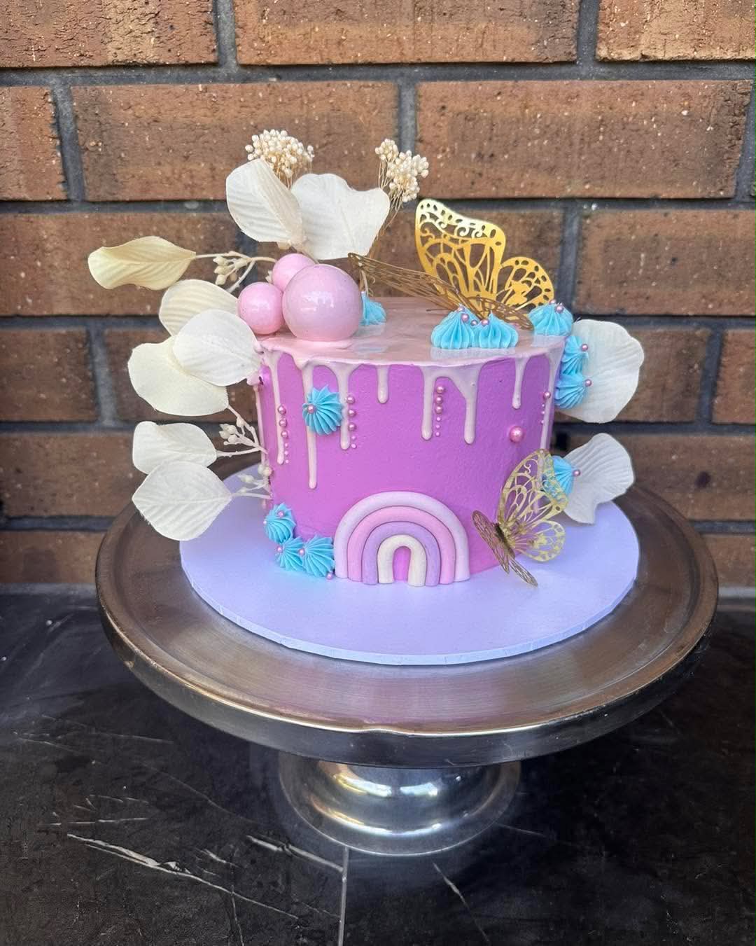Purple drip birthday cake with pink rainbow fondant, gold butterfly toppers, blue rosettes, pink sphere balls and dried white flowers